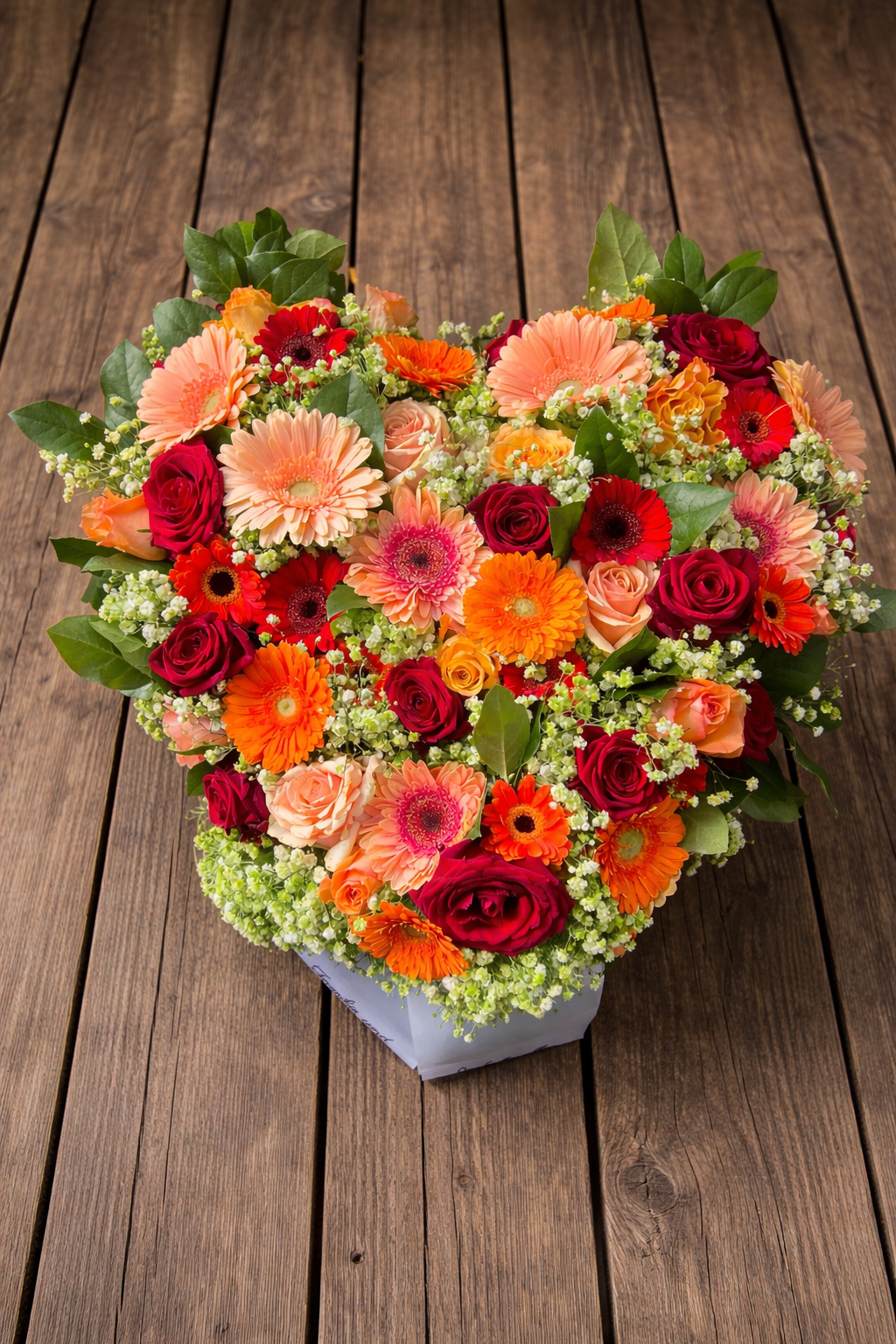 Plantula Amedorf - Trauerfloristik Herzförmiger Blumenstrauß mit roten, orangefarbenen und rosa Blüten auf einem Holztisch.
