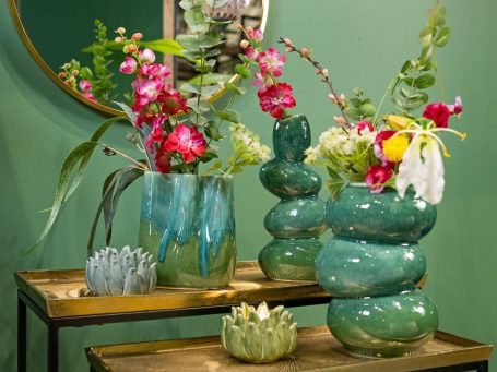 Bunte Blumenarrangements in farbigen Vasen auf einem Holztisch vor grünem Hintergrund.